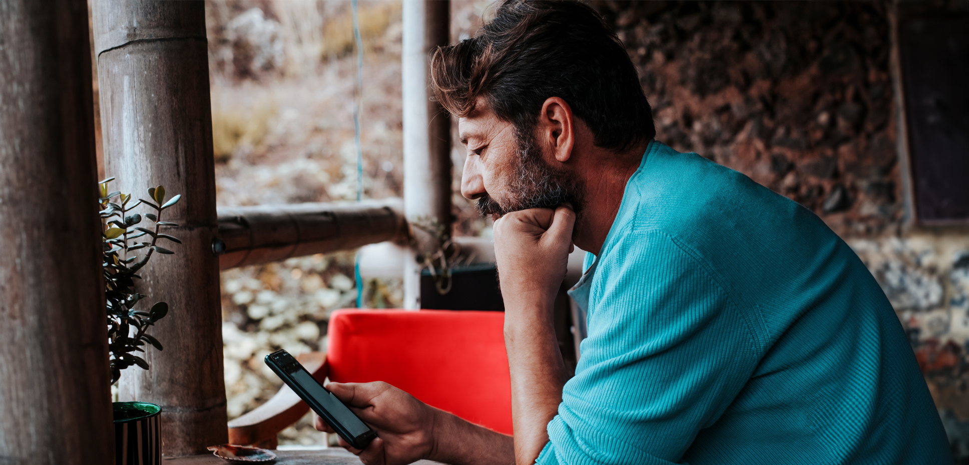 A man with brown hair and a beard looks at his mobile phone while sitting in a porch